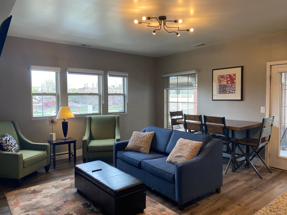 The spacious living room features a comfortable blue sofa with decorative pillows, flanked by two green armchairs. A dining area with a wooden table and metal chairs is seen in the background. Large windows allow natural light to fill the room, complemented by a modern light fixture.