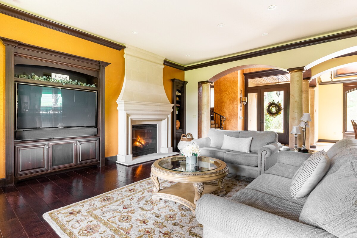 A spacious living room features a large fireplace with a stone surround and a flat-screen television mounted above a wooden console. Comfortable grey sofas are arranged around a round coffee table, complemented by a decorative area rug that adds warmth to the dark hardwood floor.