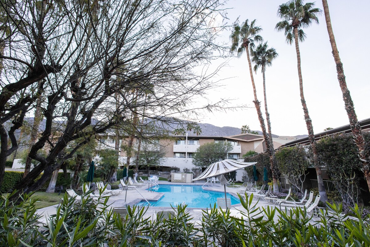 A serene pool area is framed by palm trees and lush greenery, with elegant lounge chairs positioned around the water. A shaded area offers respite from the sun, while the surrounding building features a warm, neutral facade reflecting the tranquil atmosphere.