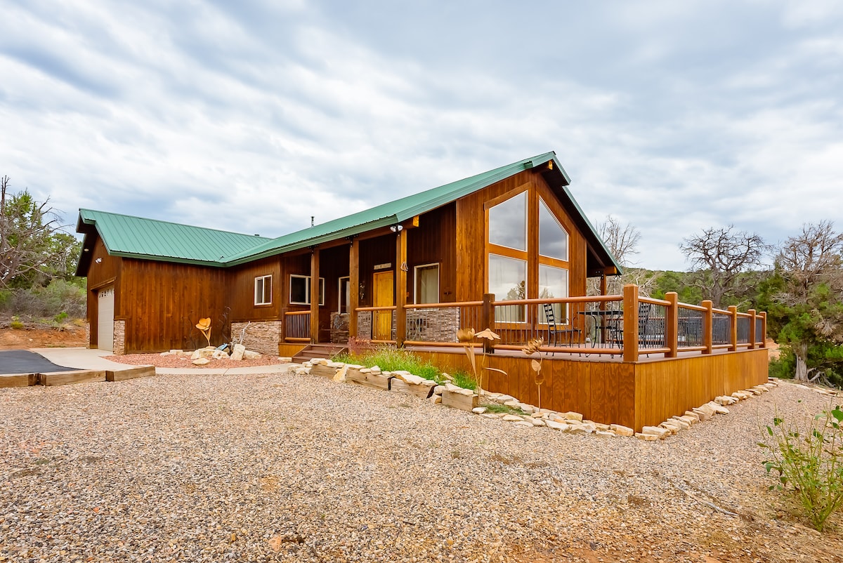 A spacious cabin is presented with a wooden exterior and a green metal roof. A large deck extends from the home, surrounded by gravel and natural landscaping. Ample windows provide views of the surrounding area, creating a connection between indoor and outdoor spaces.