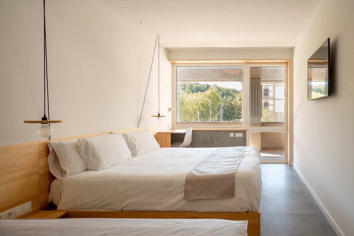 A bright and airy room features a spacious bed with neutral bedding, complemented by light wooden furnishings. A large window provides clear views of trees outside, while a flat-screen TV is mounted on the wall. A desk and chair are visible in the corner.