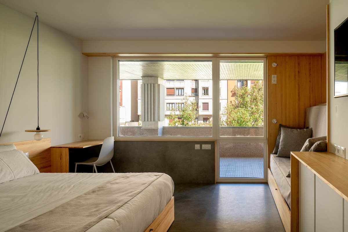 A well-lit room features a spacious bed with a neutral-colored blanket. A large window offers clear views of the surrounding trees and buildings. A desk with a chair is positioned beside the window, and a comfortable seating area is available along the wall.