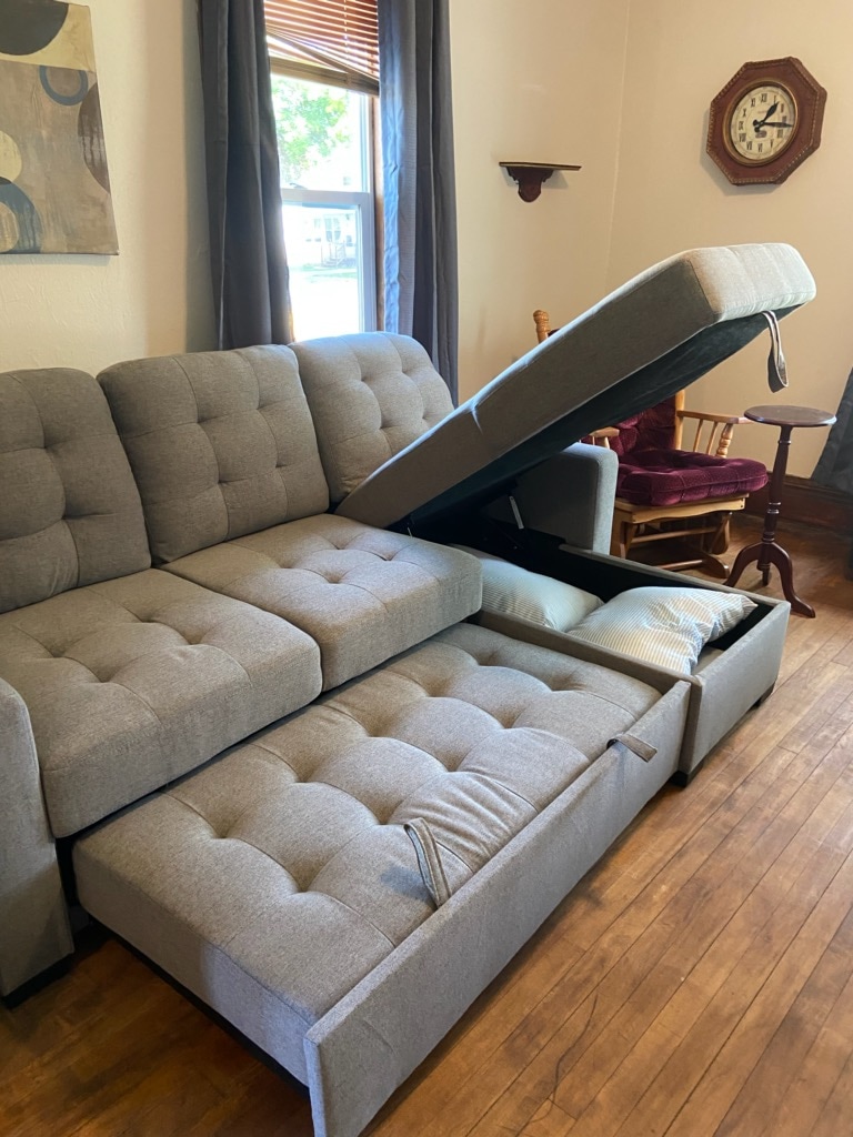 A spacious gray sectional sofa is shown with a portion of the seat lifted to reveal hidden storage underneath. Natural light enters through the nearby window, illuminating the room's wooden flooring. A wall clock and wooden chair are visible in the background.