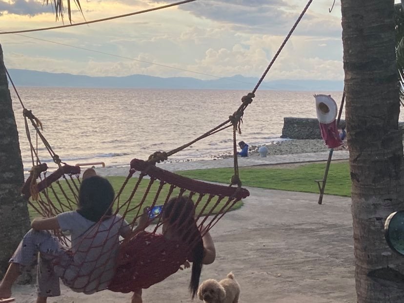 Two individuals are seated in a hammock, enjoying the view of the sea at sunset. A friendly dog is seen nearby, and a second hammock is visible hanging between two palm trees. The expansive shoreline and gentle waves contribute to a serene coastal ambiance.