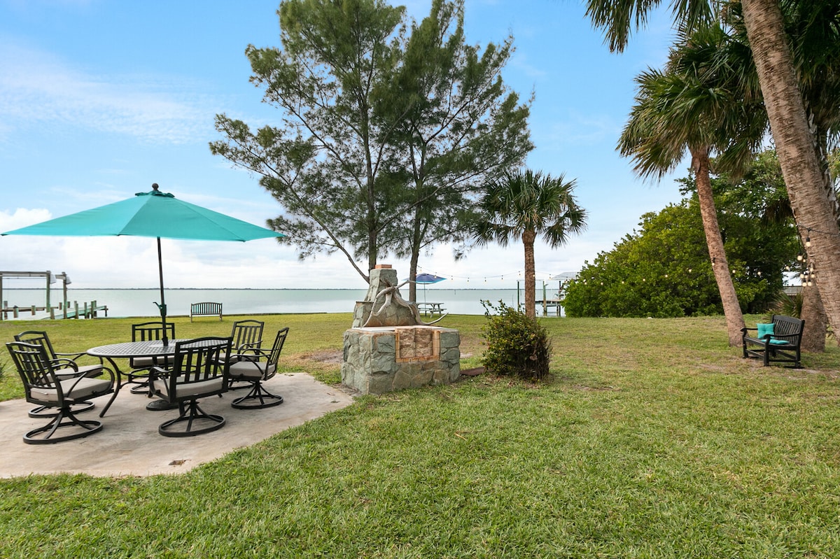 A scenic outdoor area features a stone fire pit surrounded by a circular arrangement of metal chairs. A large umbrella provides shade over a picnic table. Lush green grass and palm trees frame the landscape, with a view of the water in the background.