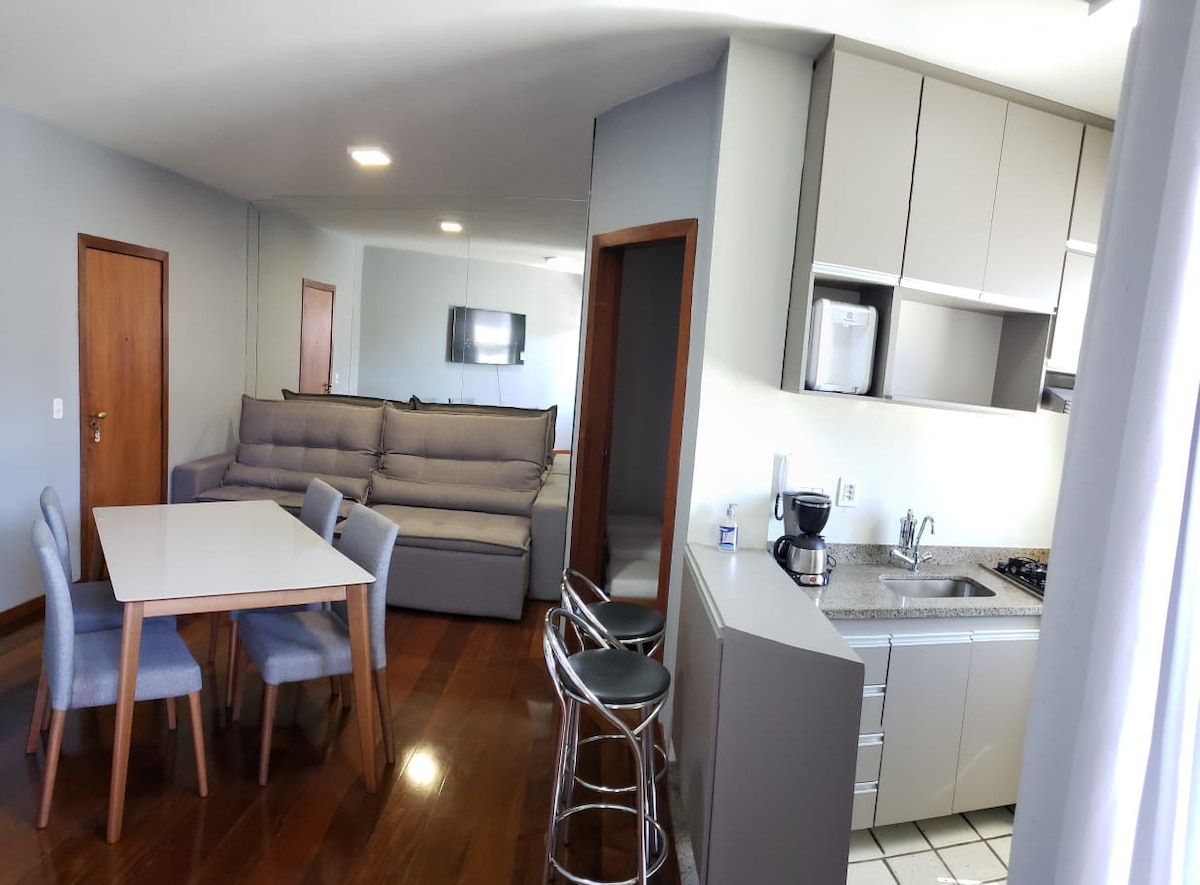 A modern living space is displayed, featuring a light-toned dining table with four chairs and a comfortable sofa positioned nearby. The kitchen area includes sleek cabinetry and appliances, while a flat-screen TV can be seen on the wall, reflecting the spacious and well-lit environment.