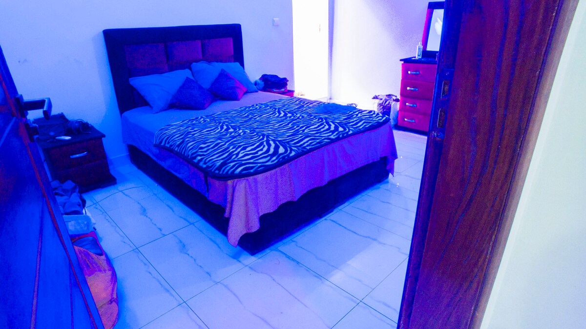 A spacious sleeping area is furnished with a large bed featuring a zebra-striped blanket. Decorative pillows are arranged on the bed, and natural light enters through a nearby doorway. A dresser with drawers is visible alongside the bed, contributing to the room's functionality.