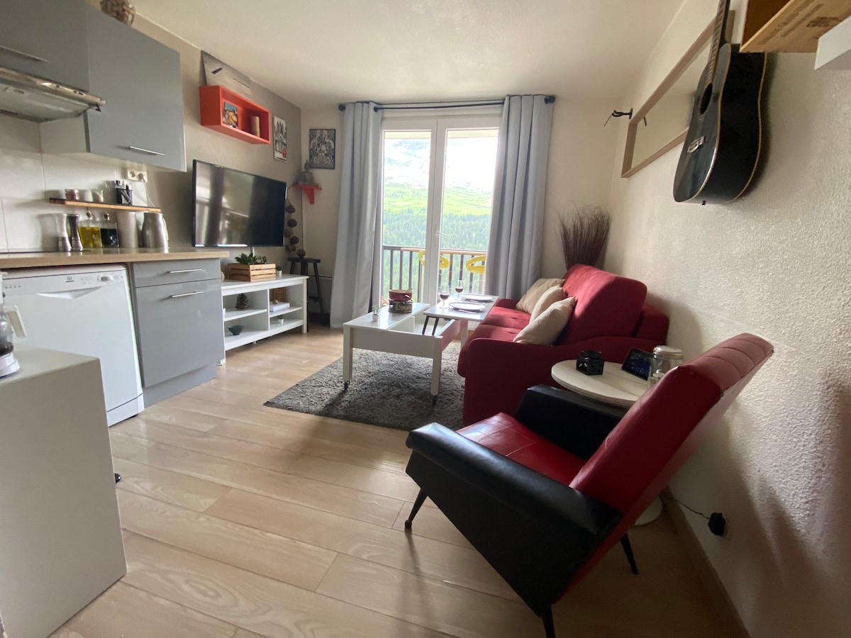 The living area displays a compact kitchen with essential appliances and a television. A red sofa and a black reclining armchair are positioned around a light rug. Natural light enters from the sliding glass doors that open onto a balcony, offering scenic mountain views.