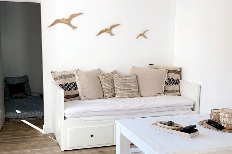 A cozy seating area features a white daybed adorned with cushions in neutral tones. Three decorative wooden birds are mounted on the wall above, complementing the light, airy ambiance of the space. A simple white table stands nearby, offering a functional touch.
