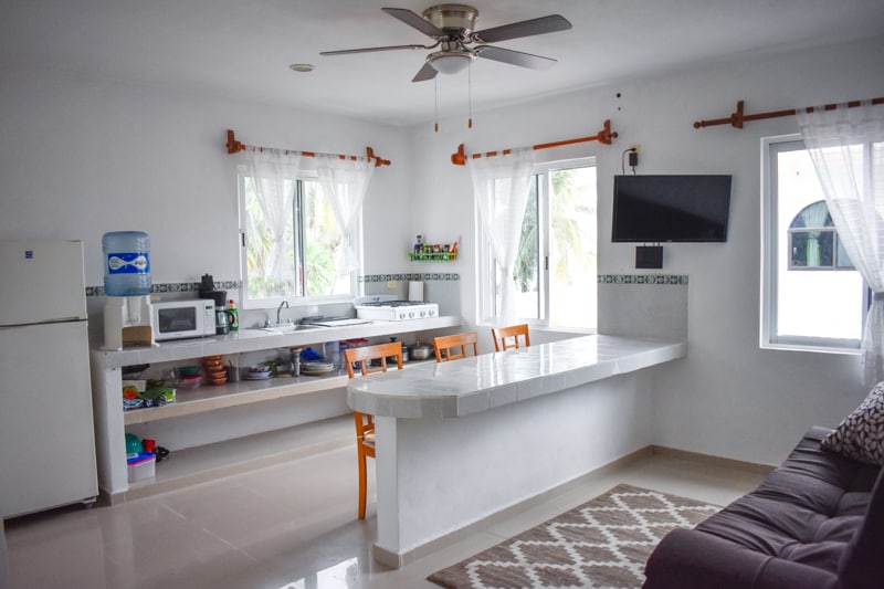 A functional kitchen area displays white cabinetry and a countertop, accompanied by modern appliances including a refrigerator, microwave, and stove. Light floods in through two windows, which are framed by white curtains. A small dining table with four orange chairs is positioned adjacent to a comfortable sofa.