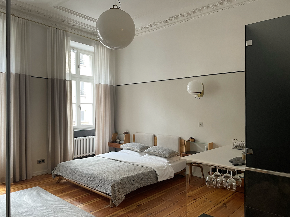 A spacious bedroom is displayed with a light-colored bed featuring neatly arranged bedding. Large windows are adorned with soft curtains, allowing natural light to fill the room. A minimalist table is visible, along with a set of hanging glassware.