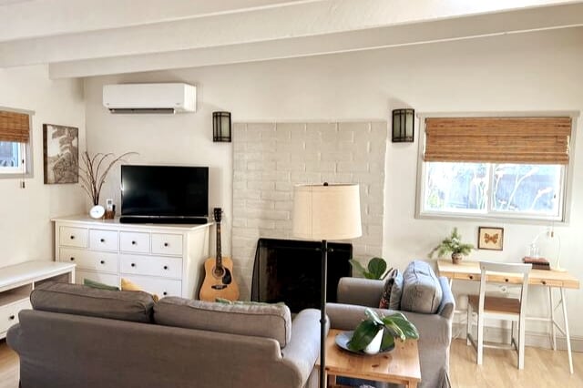 A cozy living area is presented, featuring a gray sofa and a light wooden coffee table. A television is mounted on a white cabinet beneath a decorative plant. A desk with a chair is positioned near a window, while a guitar rests against the wall beside the fireplace.