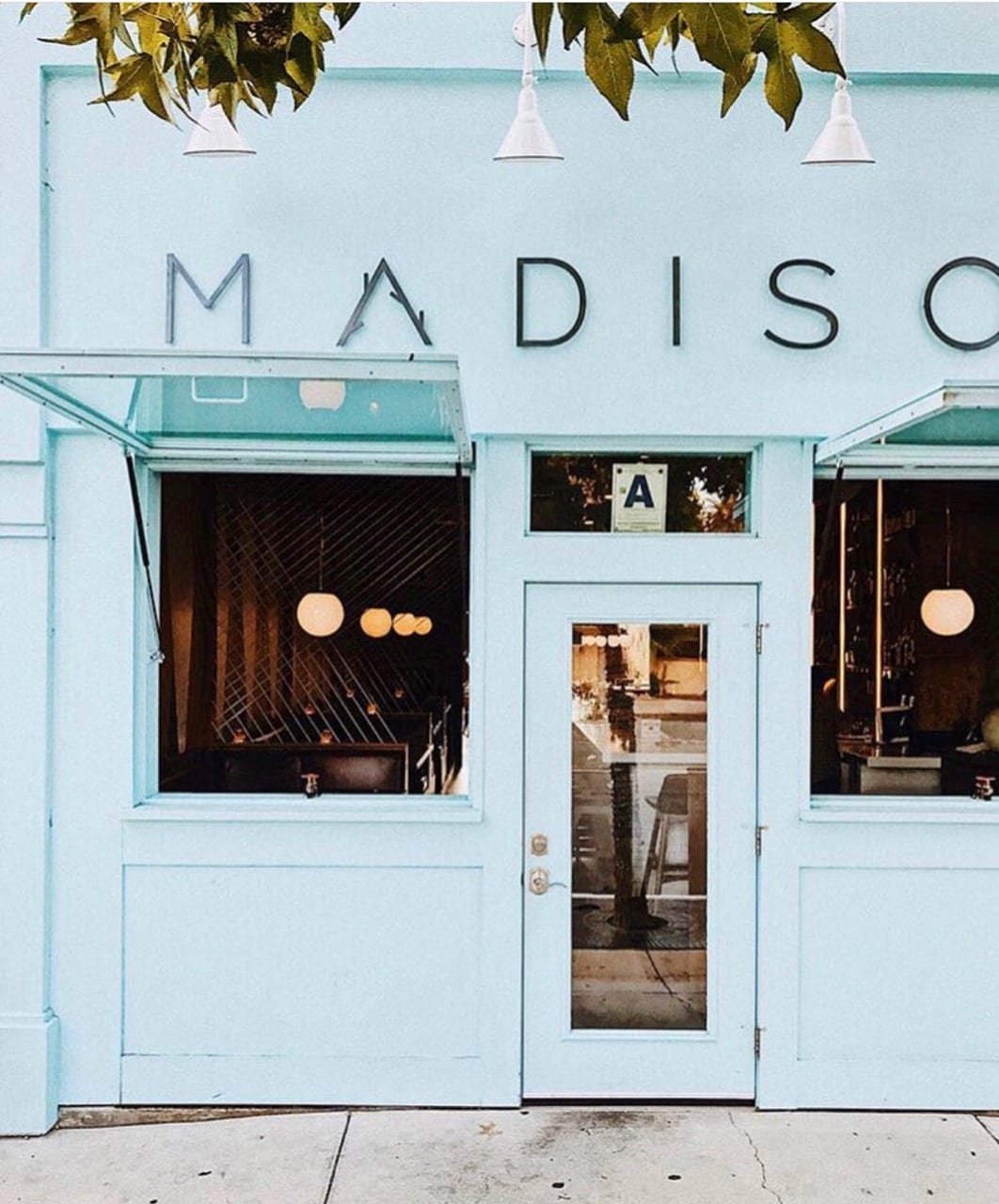 An inviting storefront is presented in a soft blue hue with large windows and a welcoming door. Round lights inside create a warm ambiance. The name 'MADISON' is prominently displayed above the entrance, enhancing the vibrant appearance of the establishment.