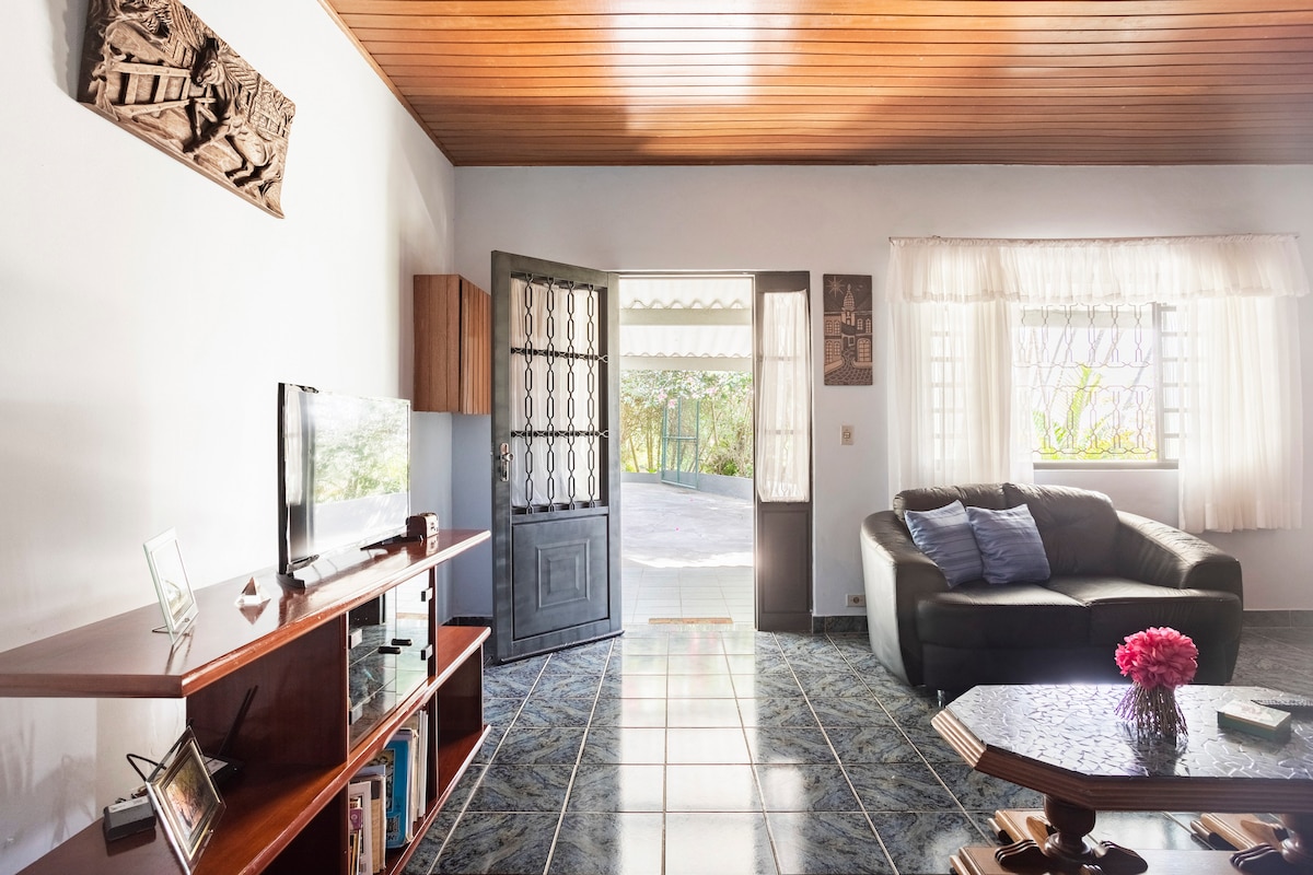 The living area showcases a dark sofa accompanied by a coffee table. A television unit is positioned to the left, displaying decorative items. Large, open doors lead to an outdoor space, while natural light floods through the windows, illuminating the room's neutral color palette.