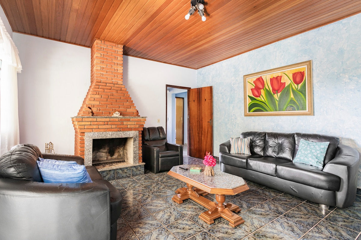 A cozy living room features a brick fireplace at its center, complemented by two black leather sofas. A wooden coffee table sits in front, adorned with a small floral arrangement. Light blue walls are accented by a colorful floral painting, creating a welcoming atmosphere.