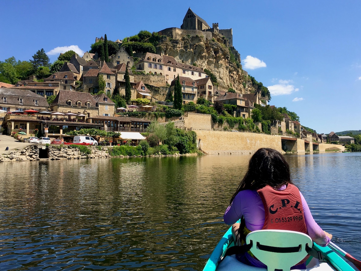 A view of the village of Beynac is captured from the water, showcasing its golden-stone houses rising against a dramatic cliff. The 12th-century château commands attention at the summit, while reflections shimmer on the calm water below, creating a serene and picturesque scene.