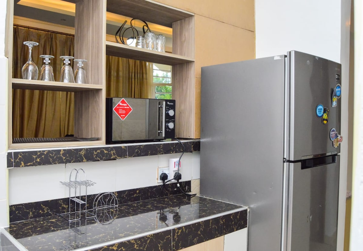 The kitchen area features a stylish dark countertop with a fitted refrigerator and a microwave. Above the countertop, shelves display glassware, while a mirror reflects the space. Warm curtains frame a window, contributing to a bright ambiance.