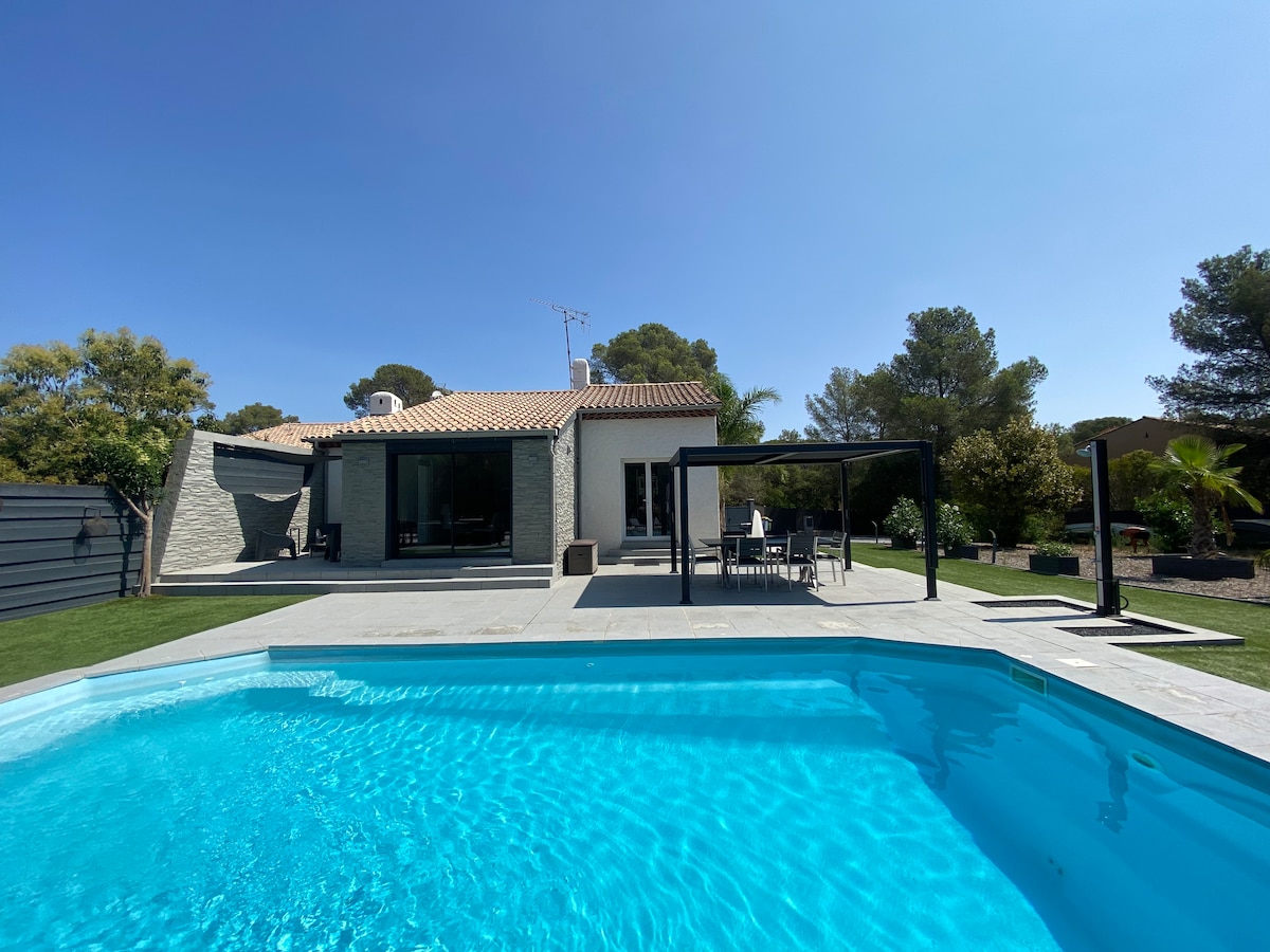 A clear view of the villa from the pool area showcases the private swimming pool with a vibrant blue surface. The exterior features a neutral color palette with a covered dining area and landscaping with greenery, creating a relaxing outdoor space.
