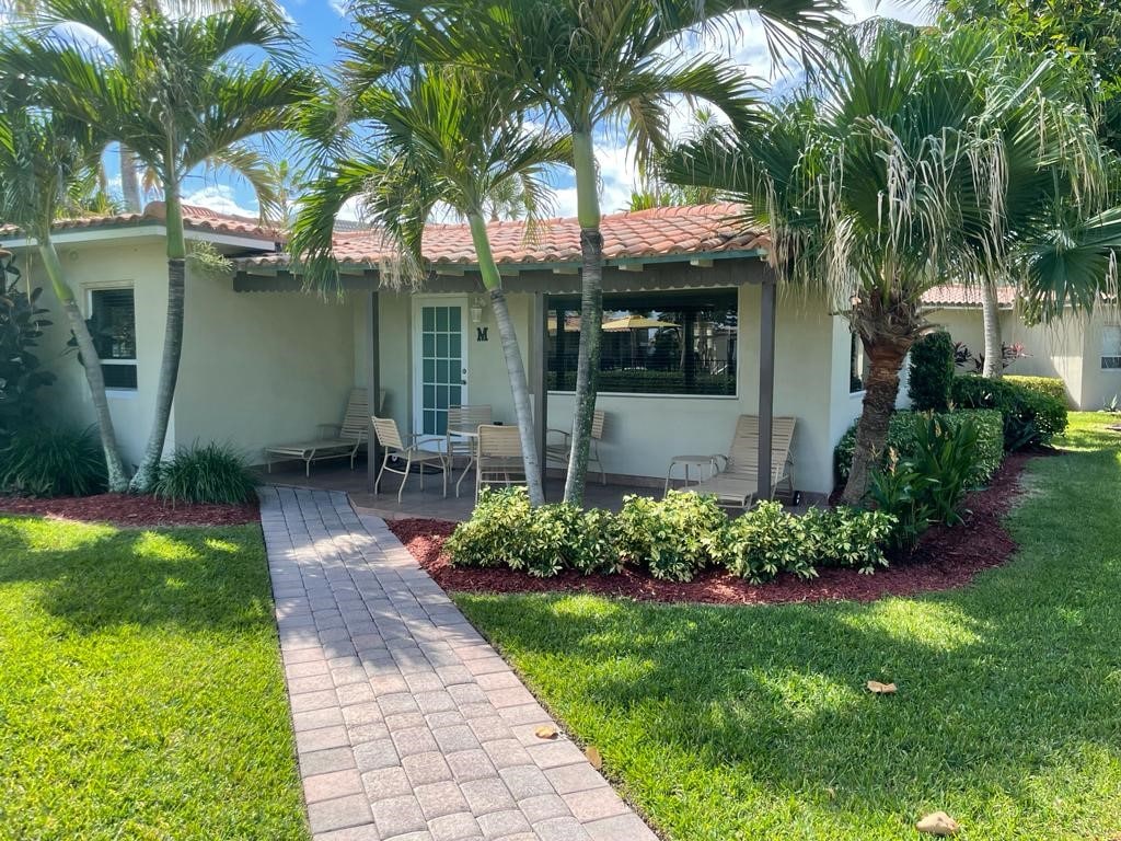 A well-maintained villa is surrounded by lush greenery and palm trees. A stone pathway leads to the entrance, framed by neatly trimmed shrubbery and colorful plants. A small seating area with light-colored chairs is positioned on the patio, inviting relaxation.