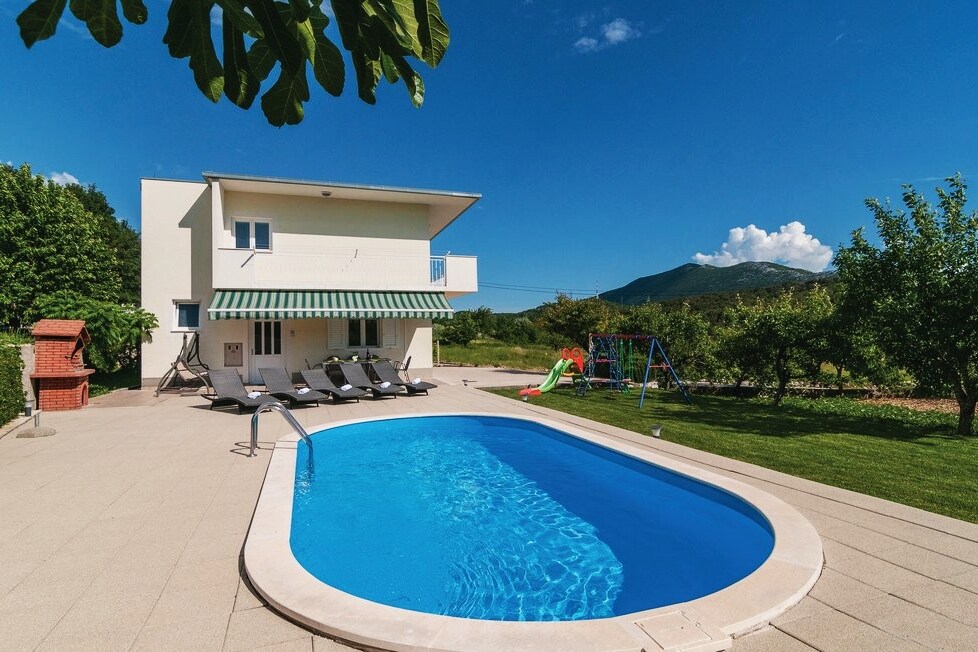 An inviting outdoor space features a private swimming pool surrounded by lounge chairs. A barbecue area is present near the house, alongside a children's playground equipped with a slide and swing. Lush greenery and distant mountains enhance the serene environment.