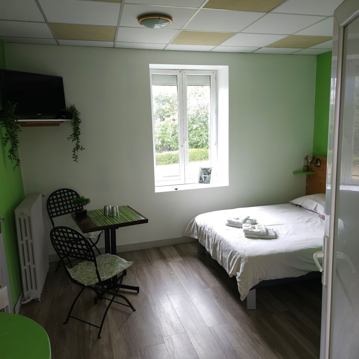 A guest room is shown featuring a spacious bed with neatly arranged linens. A small table with two chairs is placed near a window, which provides natural light. A flat-screen TV is mounted on the wall above a shelf, creating a functional space.
