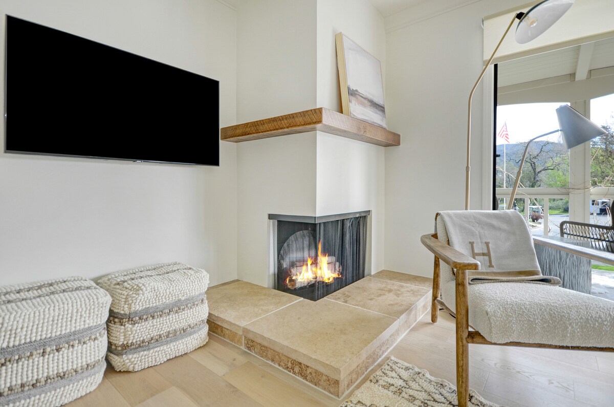 A modern living space is featured with a cozy fireplace set against a light-colored wall. A wall-mounted TV is positioned above a wooden shelf. Natural light is welcomed through a nearby window, illuminating a comfortable armchair and textured poufs on the wooden floor.