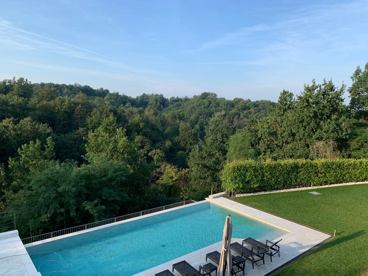 A tranquil swimming pool is surrounded by lush greenery and expansive trees. Lounge chairs are arranged around the pool, with a shaded umbrella positioned nearby. The landscape offers a scenic view of rolling hills and a vibrant natural setting.