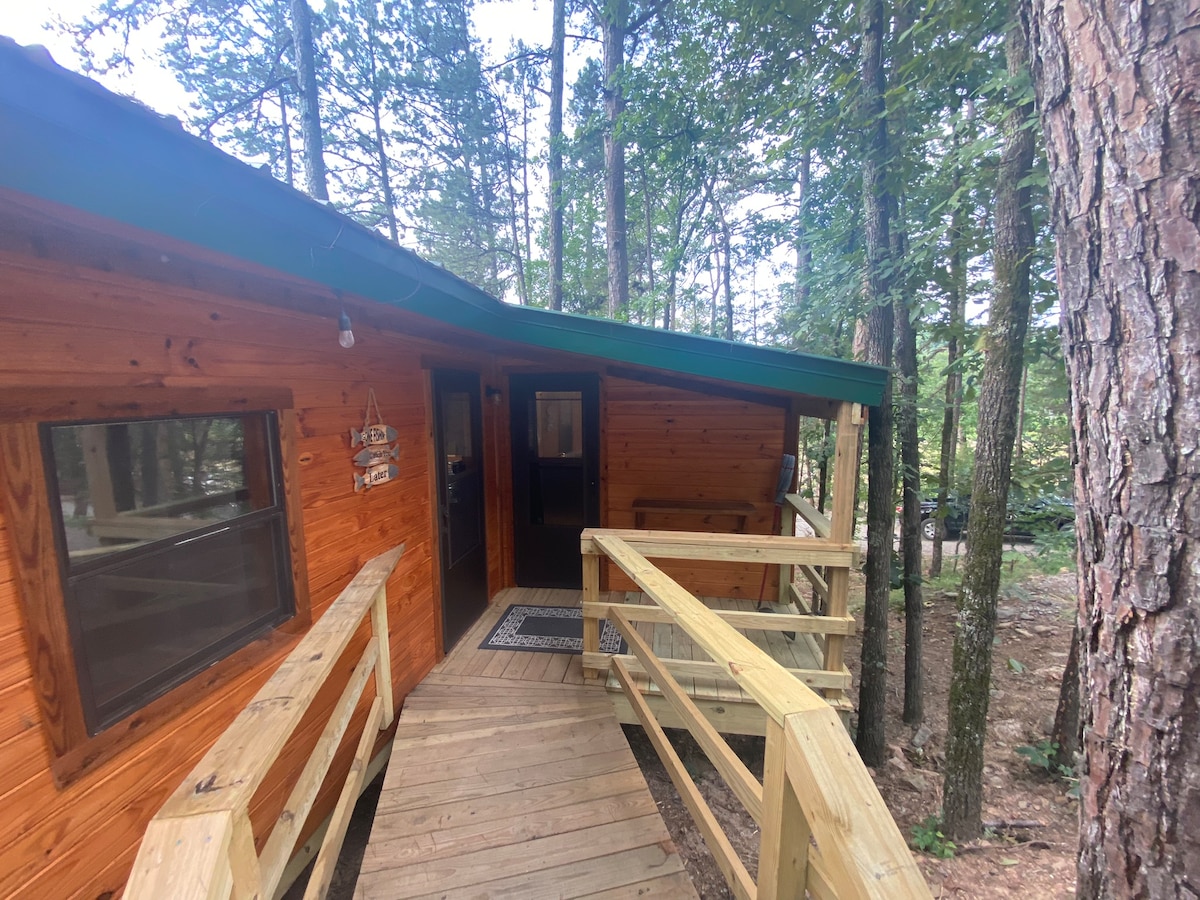 A welcoming cabin exterior is shown, featuring rich wood paneling and a green roof. A wooden ramp leads to the entrance, surrounded by tall trees. Natural light enters through the windows, inviting a peaceful atmosphere in the forest setting.
