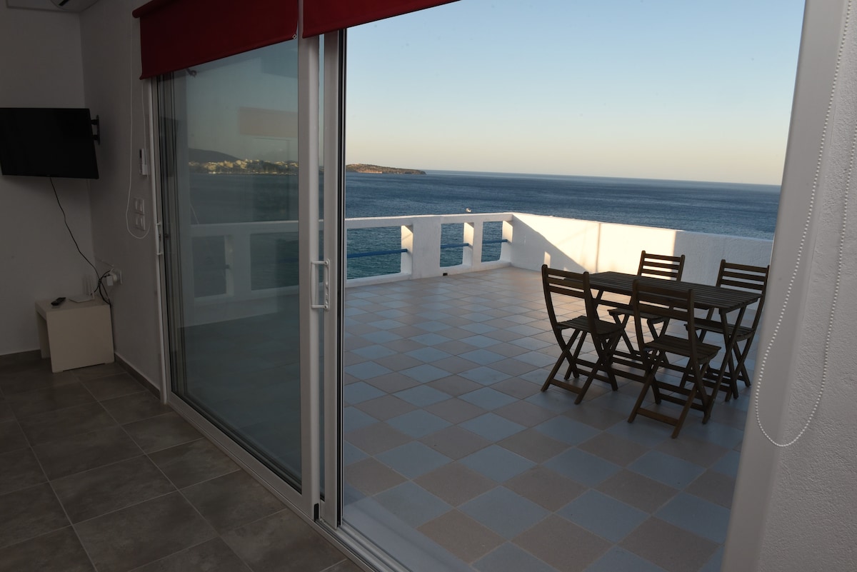 An expansive terrace is showcased, furnished with a wooden dining table and folding chairs, overlooking the serene sea view of Mirabello Gulf. Large sliding glass doors allow natural light to fill the interior space, while the calm waters create a peaceful backdrop.