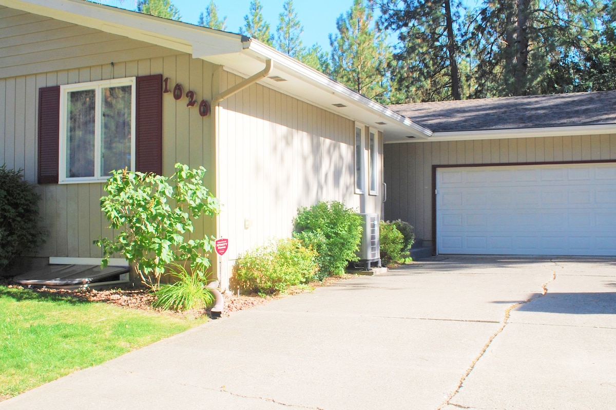 The exterior view of the home features beige siding with maroon shutters. A well-maintained driveway leads to a white garage door. Lush green shrubs and grass are visible in the front yard, surrounded by tall trees.