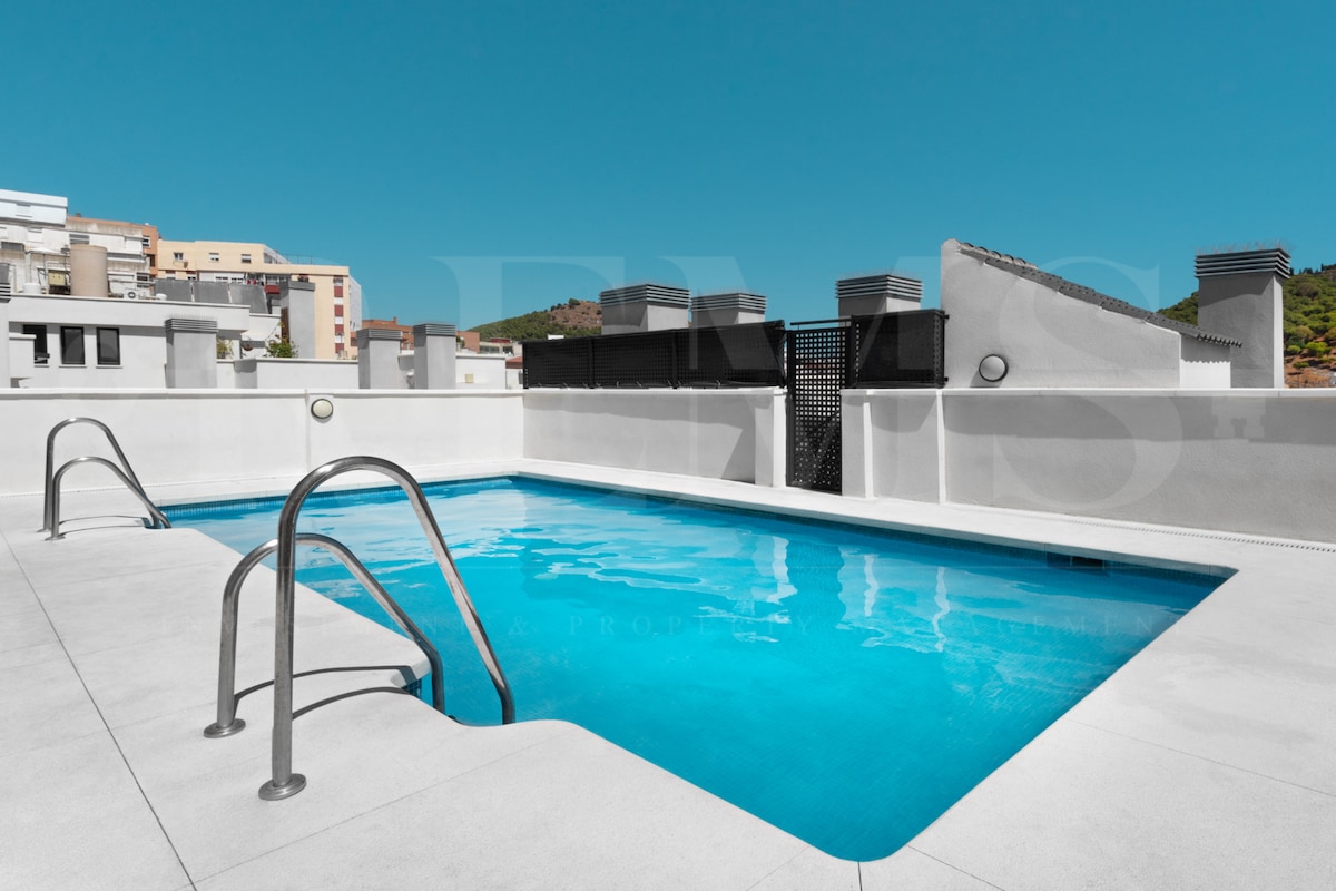A rooftop pool is featured in this image, with clear blue water inviting guests to relax. Stainless steel railing surrounds the pool area, while the surrounding terrace offers a spacious, sunlit setting, under a bright blue sky.