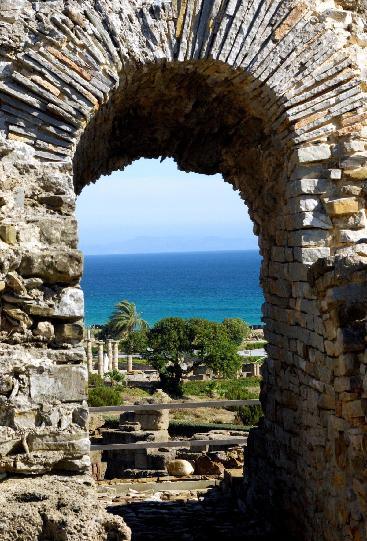 A stone archway frames a view of the coastline. The clear blue water is visible in the distance, bordered by lush greenery and tall palms. The ruins of ancient stone structures can be seen below, providing a glimpse into the historical landscape.