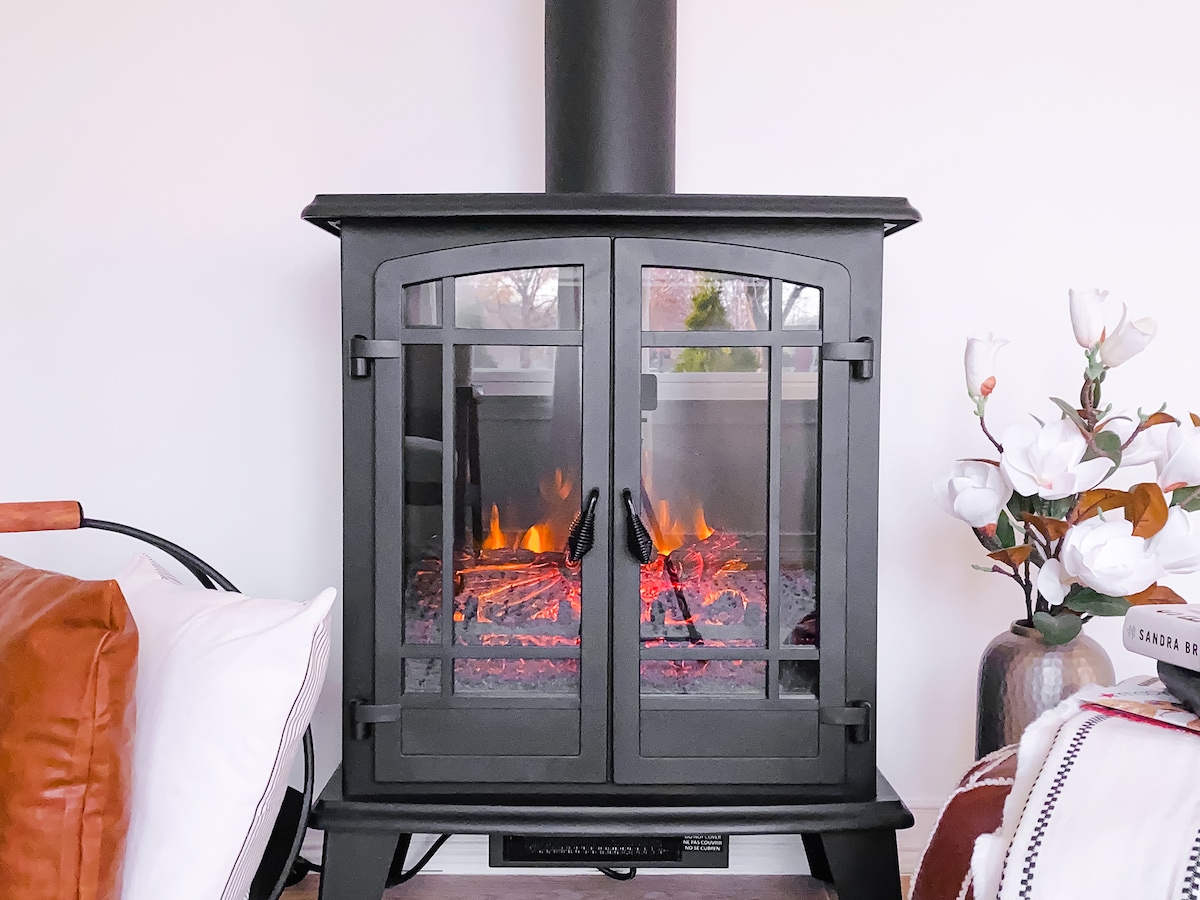 A black, modern electric fireplace features glass doors with a warm glow emanating from the simulated flames inside. Decorative white flowers in a vase and a cozy throw blanket are visible nearby, adding to the inviting atmosphere.