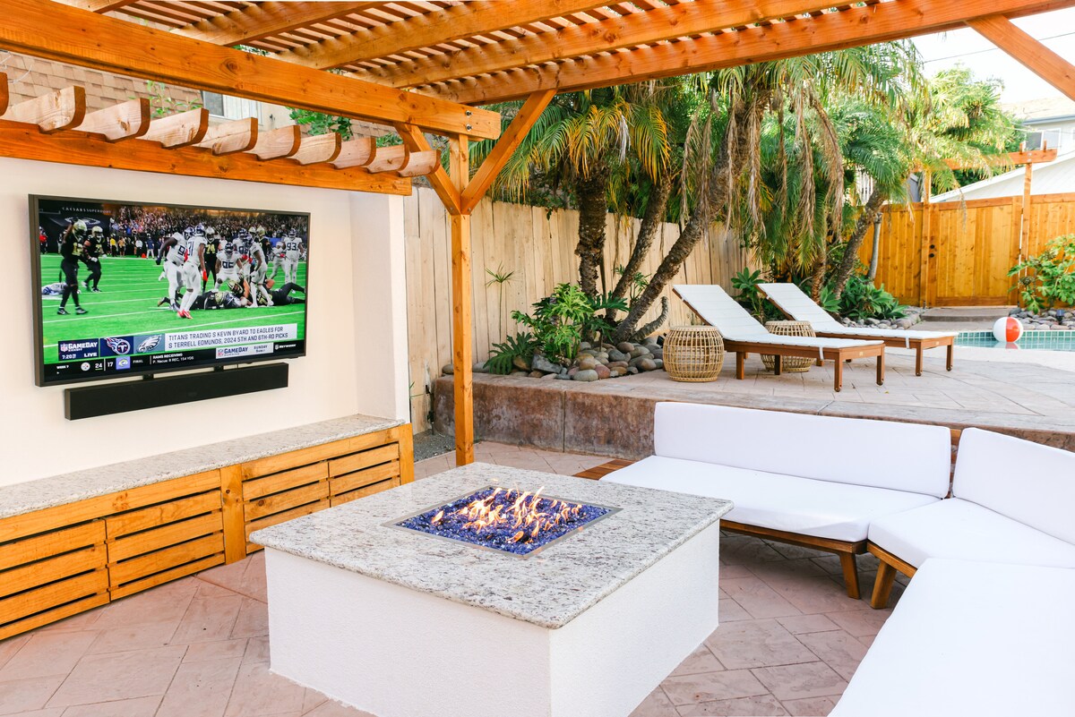An outdoor living area features a gas fire pit surrounded by a U-shaped sectional. A mounted flat-screen TV displays a live sports game nearby. The backyard is framed by lush greenery and a wooden fence, with sun loungers positioned beside a sparkling pool in the background.