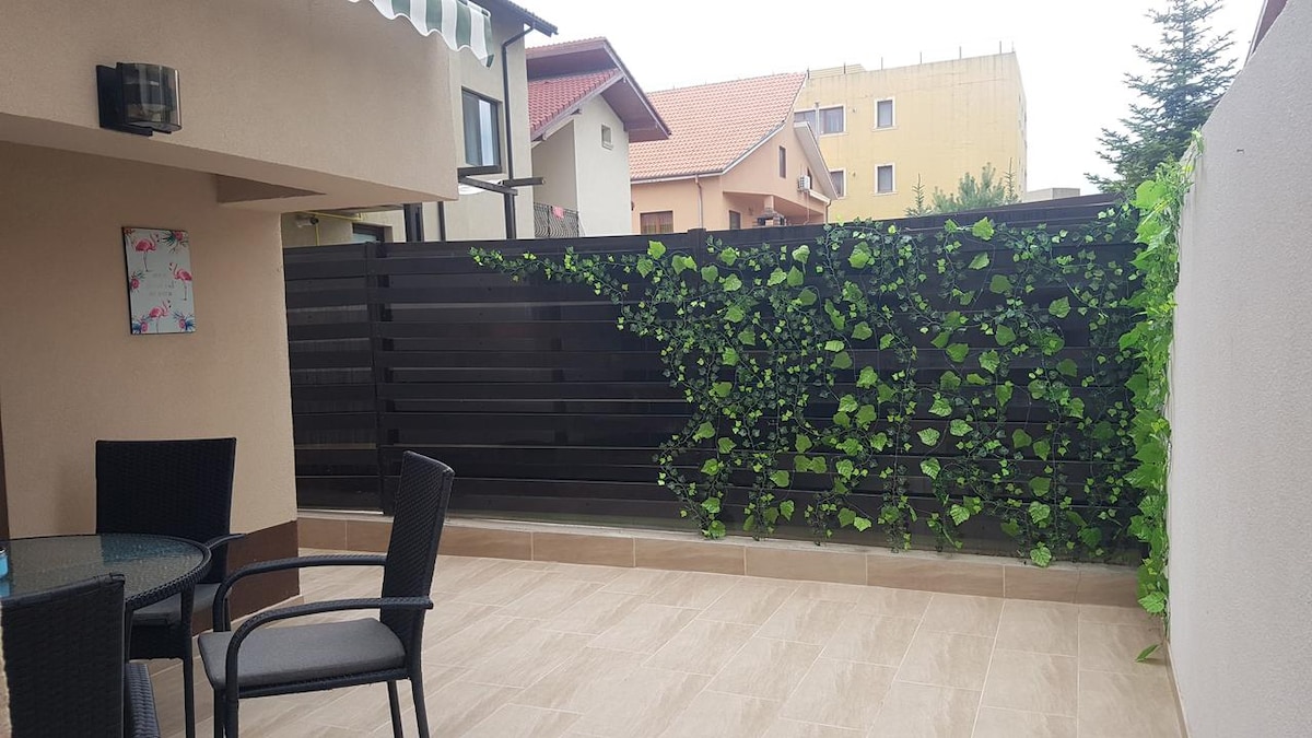 A private terrace features a sleek, dark wooden fence adorned with greenery, enhancing privacy. A round table with four black chairs is positioned on light-colored tiles, providing a comfortable outdoor space. Distant buildings and trees are visible in the background under an overcast sky.