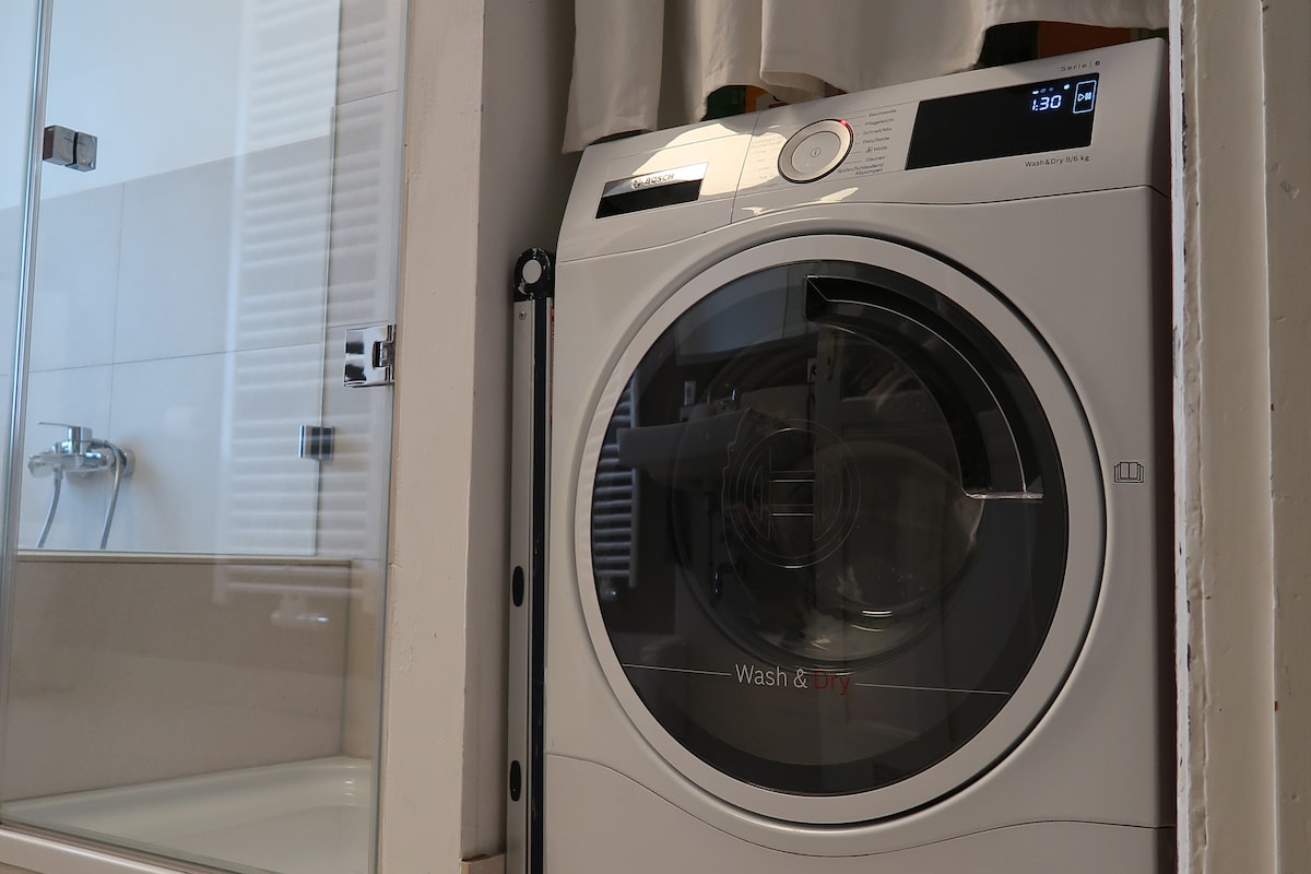A modern washing machine is positioned beside a spacious shower area, integrating functionality within the bathroom. The sleek design of the machine is visible, complemented by neutral tile tones in the background, enhancing the overall practicality of the space.