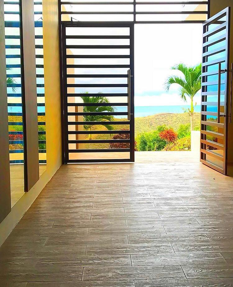 The entrance features large, modern doors that create an inviting transition between indoor and outdoor spaces. A scenic view of distant greenery and palm trees is visible through the slatted door design, with natural light illuminating the wood-toned flooring.