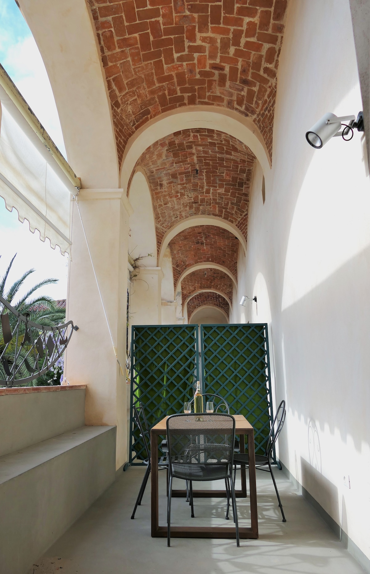 A charming outdoor space features a dining table with four chairs, framed by an arched brick ceiling. Green lattice adds a touch of greenery, while ample sunlight brightens the area, creating an inviting setting for meals or relaxation.