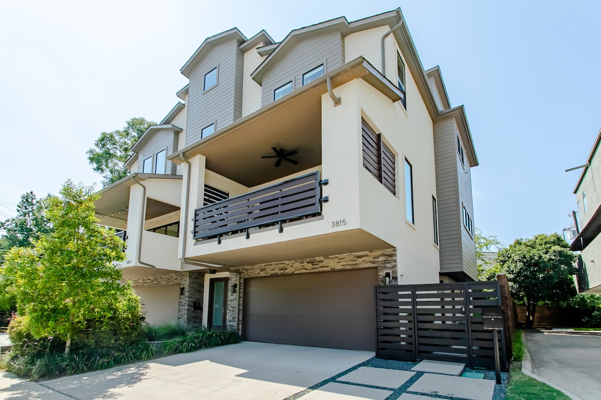 A modern four-story attached home is featured, displaying a combination of earthy tones and contemporary architecture. A spacious garage is visible, accompanied by a well-maintained driveway. The second floor includes a balcony with railings, while greenery surrounds the entrance, adding a touch of nature.