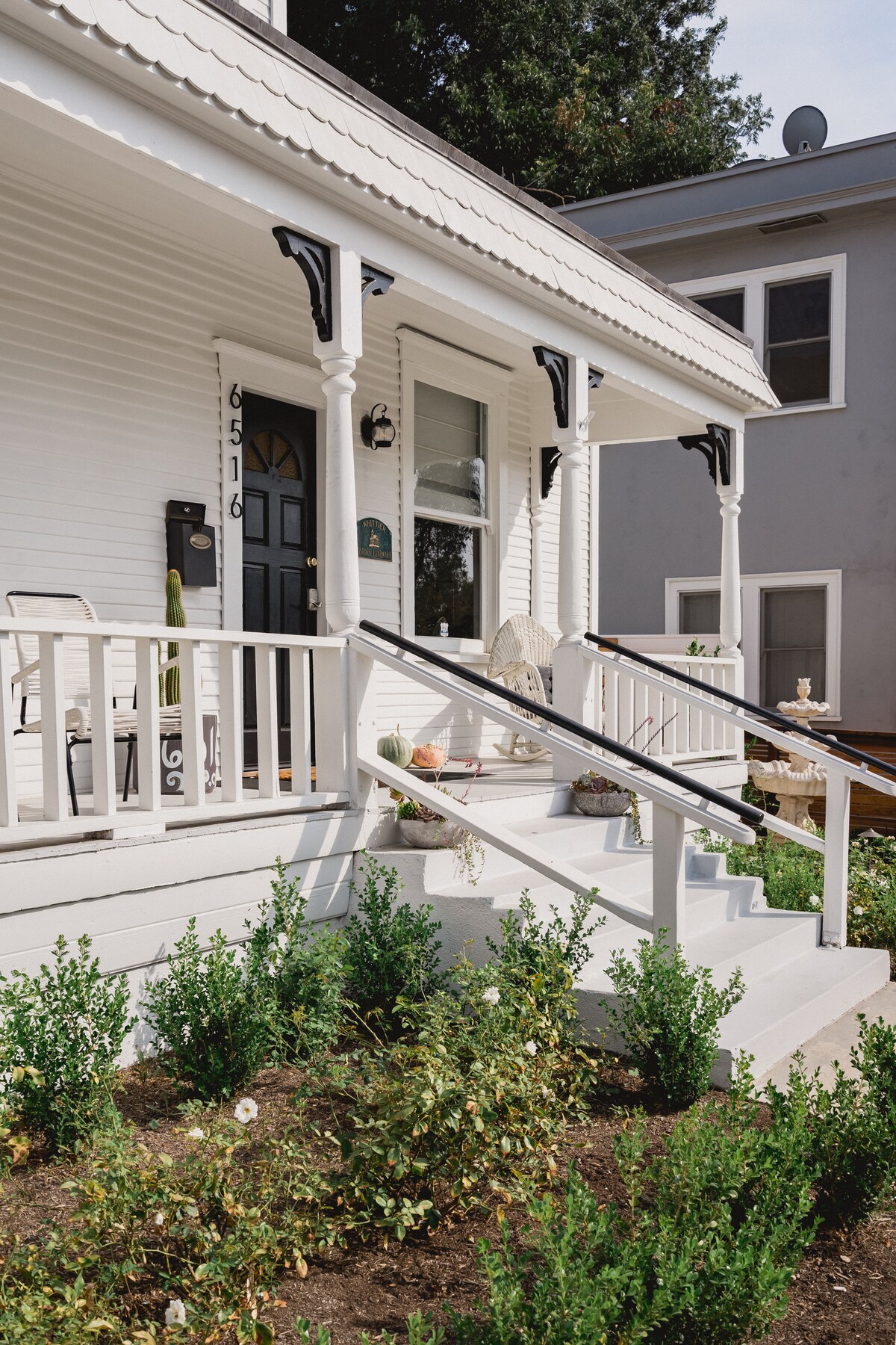 An inviting front porch features white railing and steps leading up to the historic home. Soft green plants surround the entrance, adding a touch of nature. Light streams through a nearby window, highlighting the charming architectural details of the house.