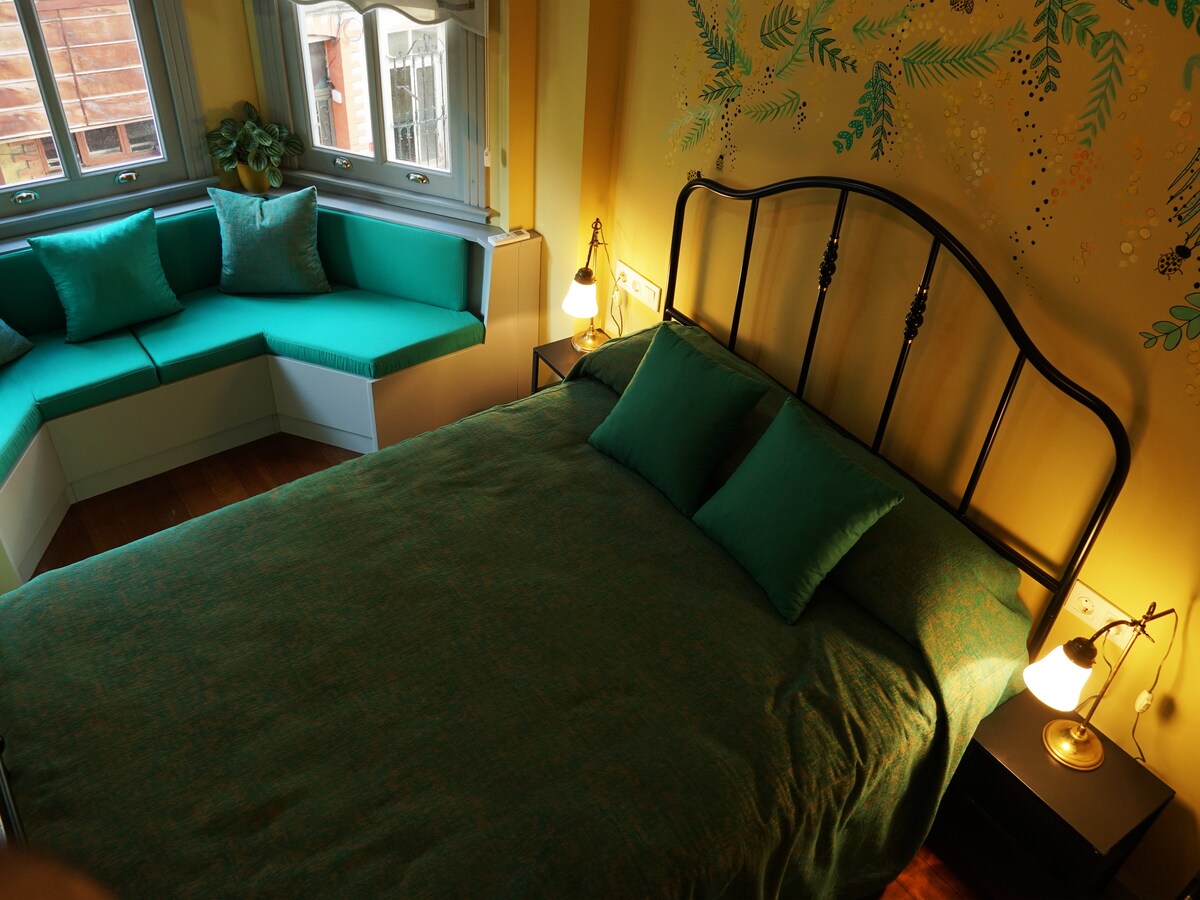A spacious bedroom features a black metal bed with teal bedding and two matching pillows. Natural light enters through large windows adorned with green seating. A decorative wall with soft illustrations complements the room's color palette.