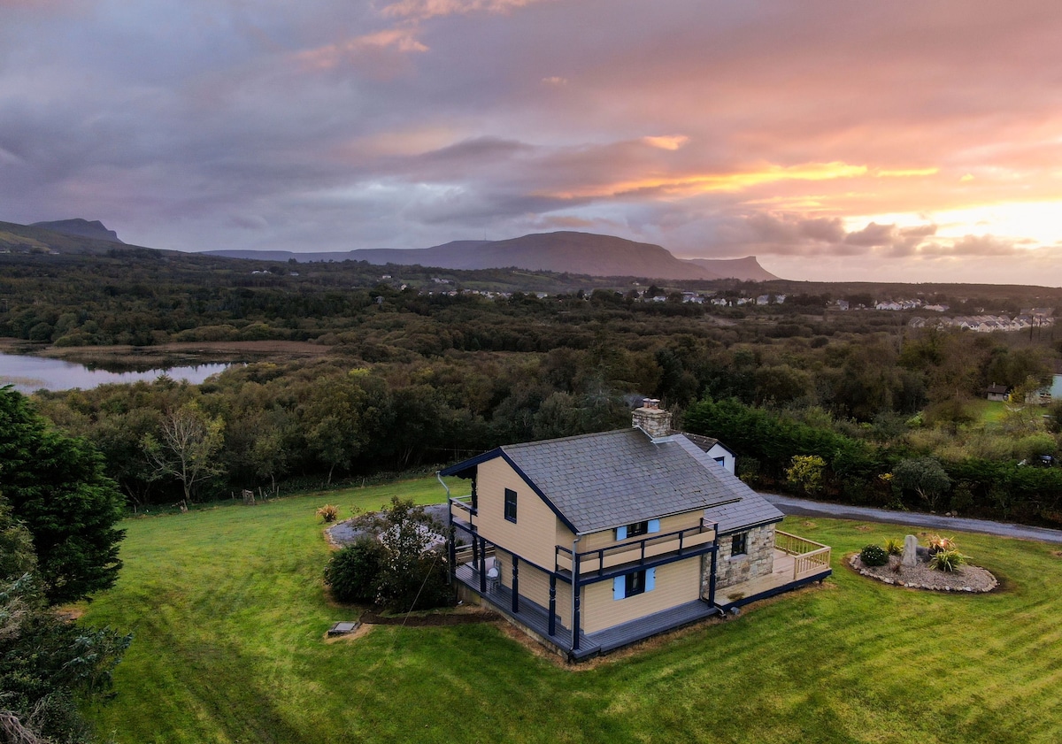 The lodge is nestled in a lush, expansive landscape, framed by rolling hills and a tranquil lake. A striking sunset casts warm tones across the sky, enriching the natural surroundings. The property features a well-maintained lawn and a charming outdoor seating area.