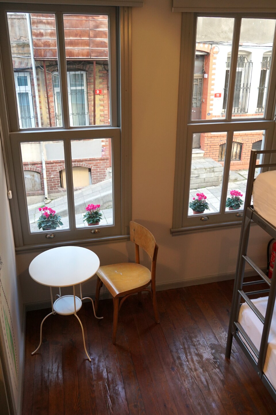 A corner of the room features two windows with gray frames, allowing natural light to illuminate the space. Below, a round white table is complemented by a wooden chair. Outside, colorful flower pots adorn the sidewalk, enhancing the view of the street.