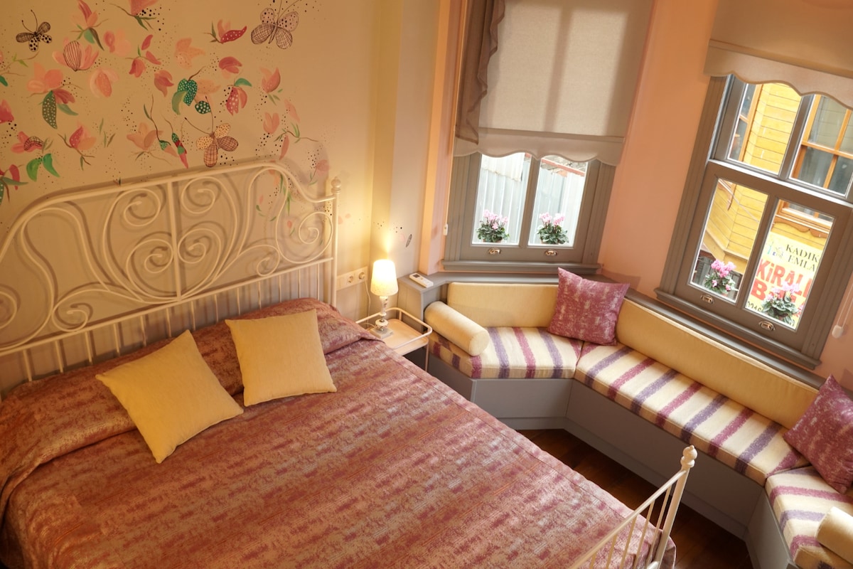 A softly lit bedroom features a spacious bed with patterned bedding and two accent pillows. A built-in seating area with cushions provides a cozy nook by the window, where floral decorations can be seen. Pastel-colored walls enhance the room's serene ambiance.