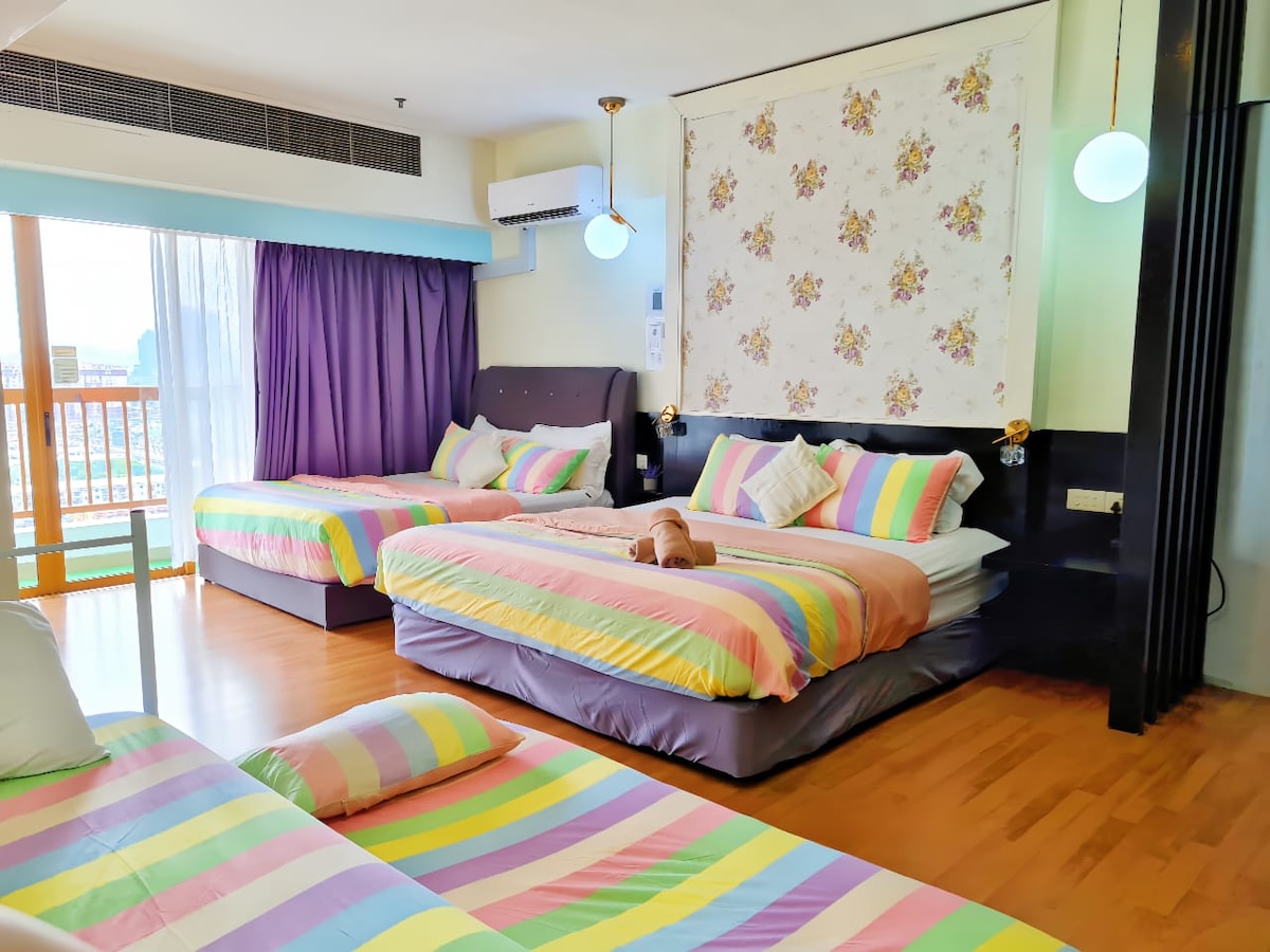 A brightly colored room features multiple beds with striped bedding in pastel hues. Soft pillows are scattered on the floor, and natural light enters through large windows lined with sheer curtains. Decorative elements include patterned wall art, adding a cheerful ambiance.