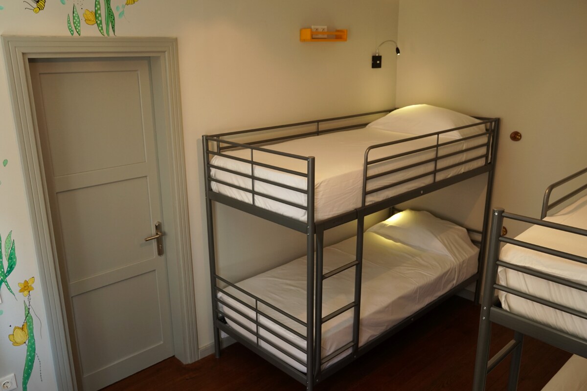 A shared sleeping area features two metal bunk beds with clean, minimalist lines. Soft lighting is positioned above the beds, illuminating the white bedding. A door is visible on the left, while colorful wall art adds a touch of character to the space.