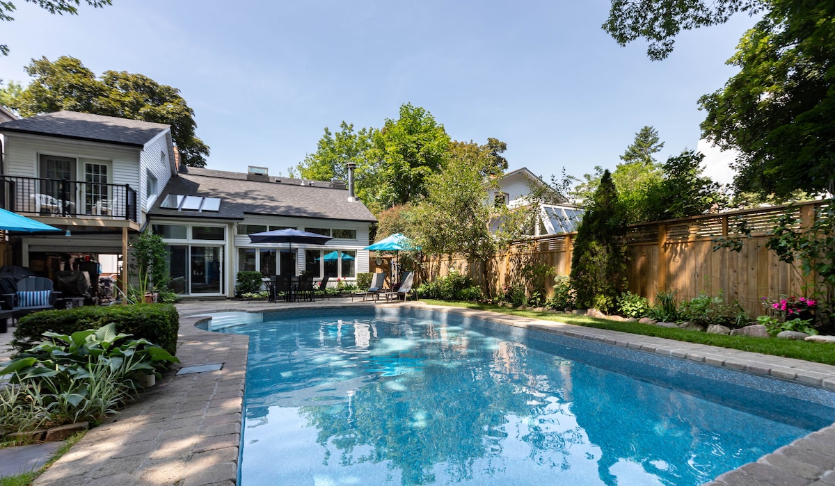 A private backyard oasis is presented, featuring an in-ground saltwater pool surrounded by lush landscaping. Comfortable lounge chairs are positioned around the pool area, and a variety of trees provide natural shade. The home is visible in the background, adding to the serene atmosphere.