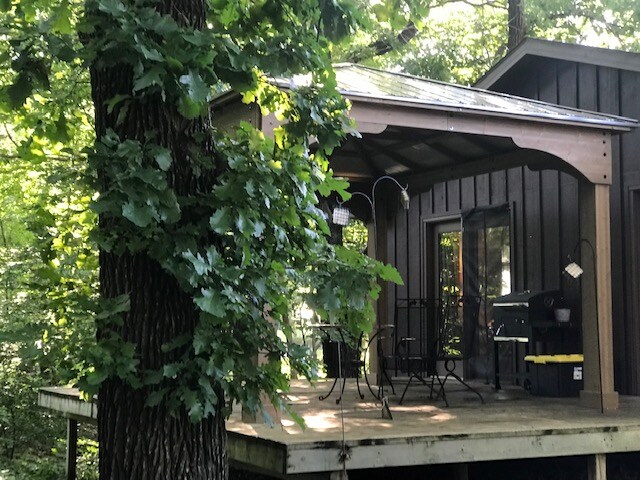 A cozy outdoor seating area is visible on the deck of a guest house, surrounded by lush greenery. A shaded pergola provides shelter, while a small table and metal chairs invite relaxation. Large trees frame the structure, enhancing the natural setting.