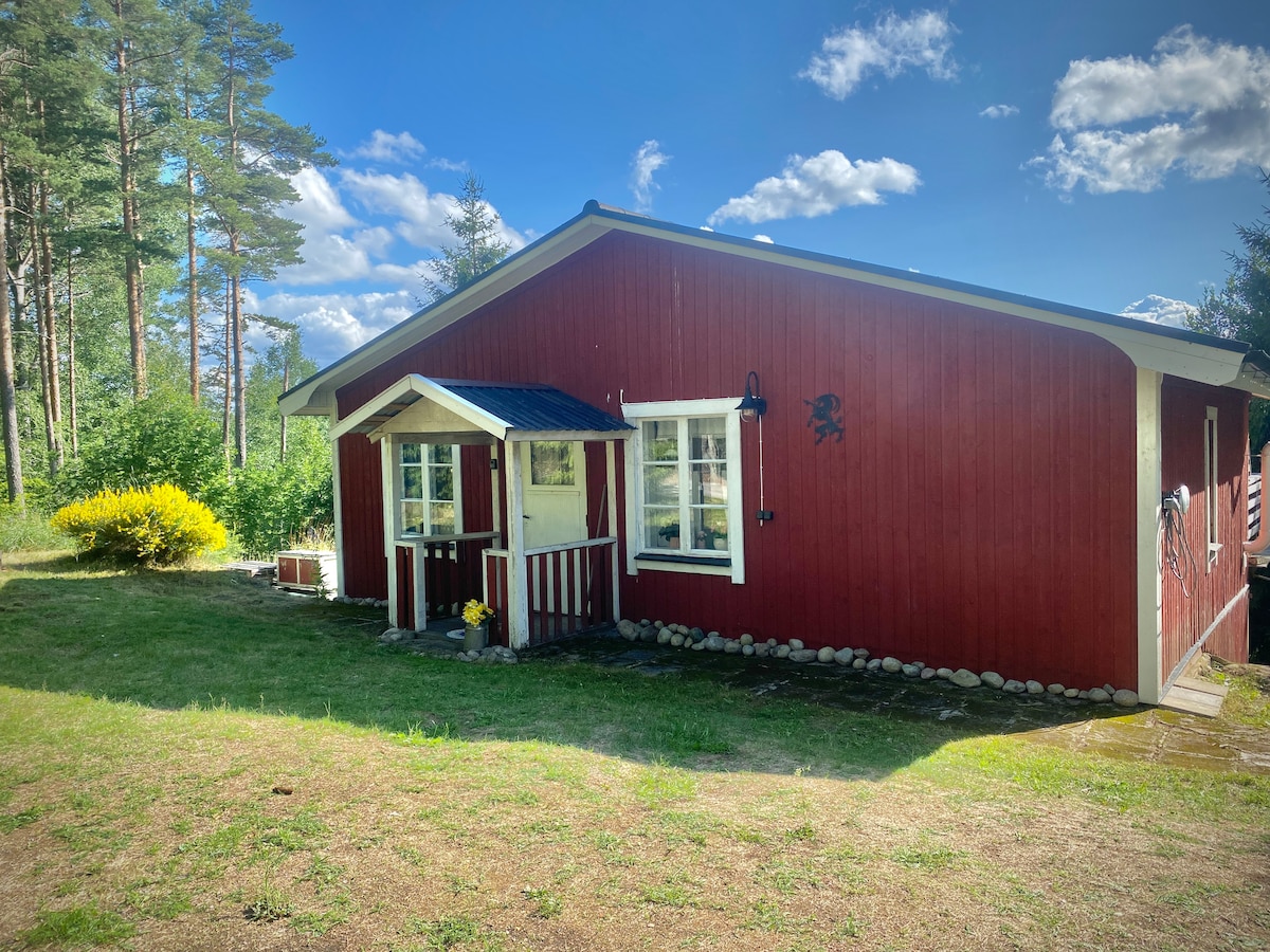A red wooden cabin is framed by lush greenery and soft grass, with bright blue skies and fluffy clouds above. The entrance features a small porch with white window frames, while garden flowers add a touch of color to the landscape.