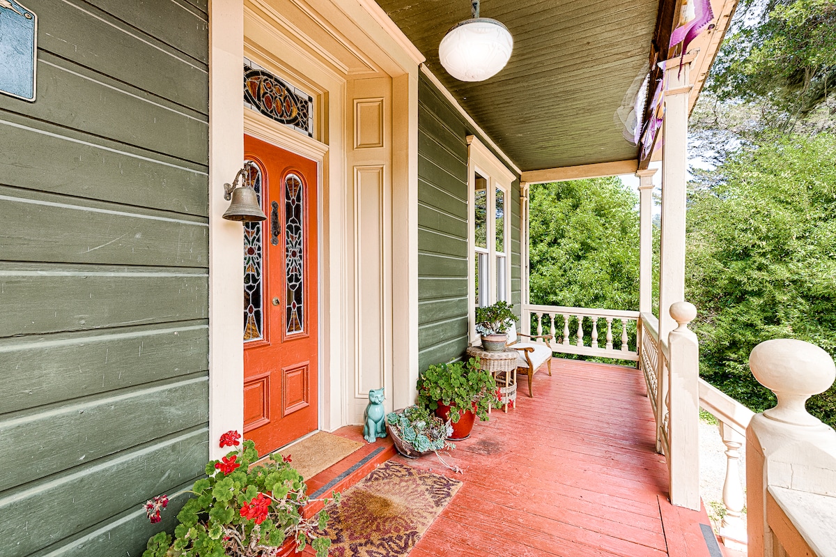 The welcoming front porch features a bright orange door framed by intricate stained glass. Potted plants and a decorative rug enhance the charming entrance, while the wooden flooring adds a rustic touch. Natural greenery is visible beyond the railings, creating a peaceful ambiance.